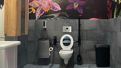 Blick in ein behindertengerechtes WC, dessen Wände oberhalb der gefliesten Fläche ein Blumenmuster auf dunklem Grund zeigen.