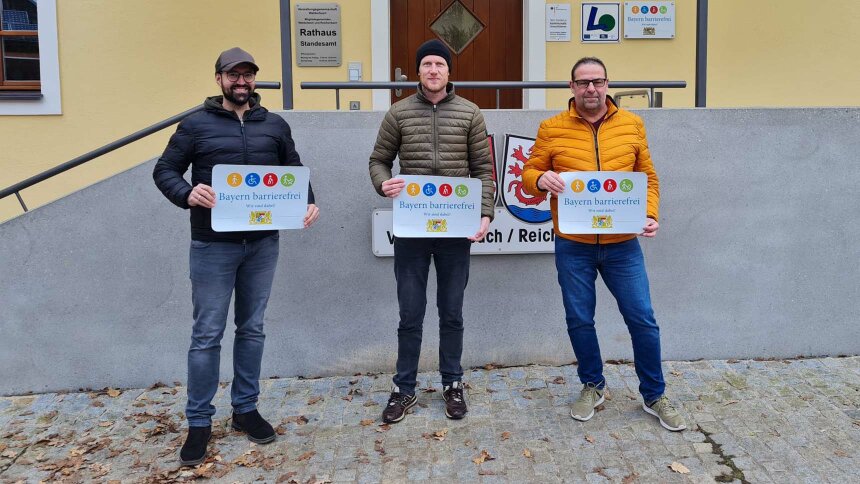 Vor dem Aufgang zu einem Gebäude: Drei Männer präsentieren das Signet „Bayern barrierefrei“.