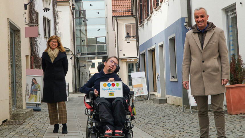 Drei Menschen, im Rollstuhl bzw. zu Fuß, mit dem Signet „Bayern barrierefrei“ unterwegs in einer Altstadtgasse.