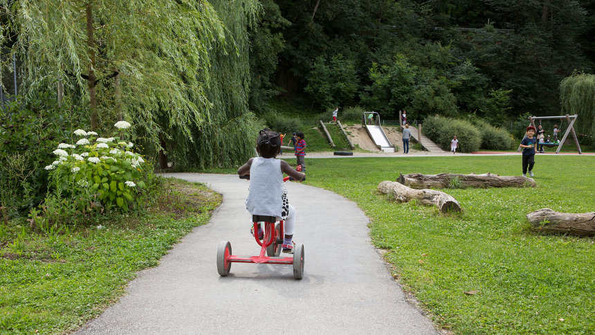 In einer Gartenanlage: Ein Kind fährt mit einem Dreirad auf einem asphaltierten Weg.