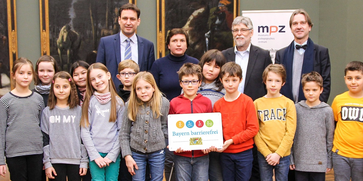 Gruppenbild: Grundschulkinder und vier Erwachsene präsentieren das Signet „Bayern barrierefrei“.