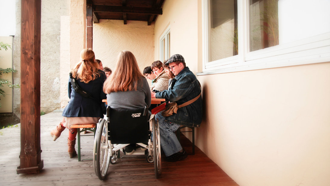 Terrasse mit Biertisch und Bänken. Eine Gruppe von Menschen sitzt um den Tisch; am Kopfende eine Frau im Rollstuhl.