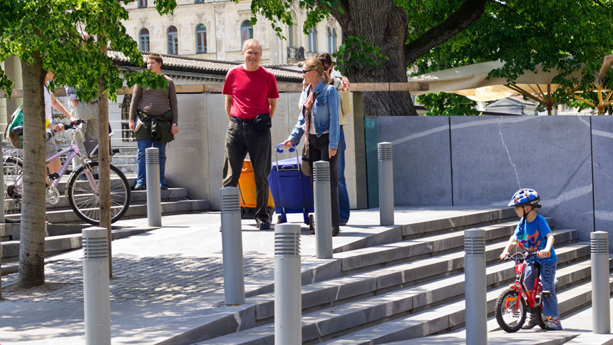 Erwachsene mit Einkaufs-Trolleys und kleines Kind mit Fahrrad auf einem öffentlichen Platz mit Stufen und Rampe.