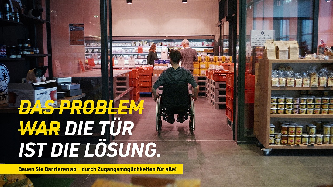 Ein Rollstuhlfahrer fährt durch eine schwellenlose Glasschiebetür in ein Geschäft. Auf dem Bild steht: „Die Tür ist die Lösung. Bauen Sie Barrieren ab – durch Zugangsmöglichkeiten für alle!“.