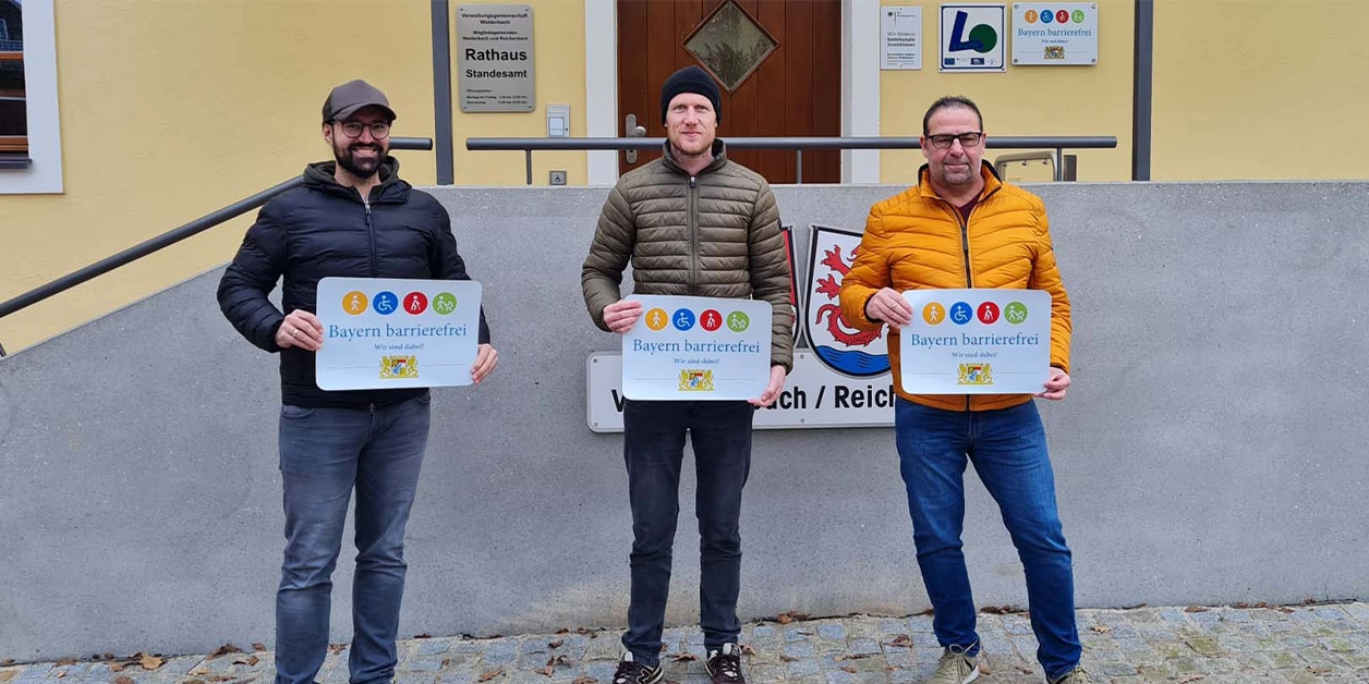 Vor dem Aufgang zu einem Gebäude: Drei Männer präsentieren das Signet „Bayern barrierefrei“