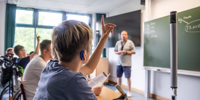 Ein Junge hebt im Schulunterricht die Hand. Er trägt ein Hörgerät. 