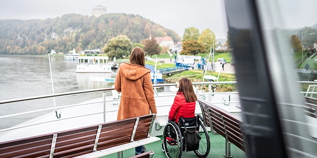 Eine Mutter steht mit ihrer Tochter im Rollstuhl auf einer Fähre.