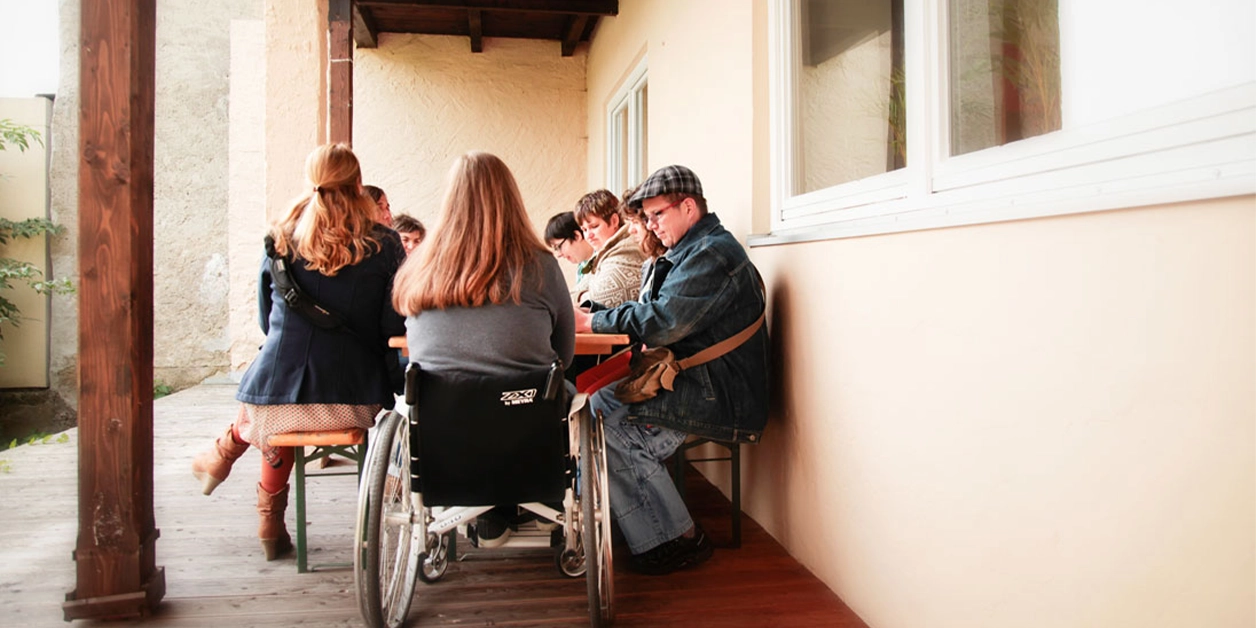 Terrasse mit Biertisch und Bänken. Eine Gruppe von Menschen sitzt um den Tisch; am Kopfende eine Frau im Rollstuhl.