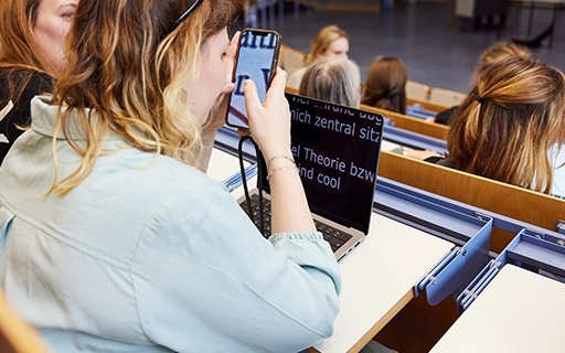 Eine Frau in einem Hörsaal. Auf ihrem Laptop ein Text in vergrößerter Schrift. 