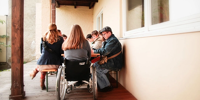 Terrasse mit Biertisch und Bänken. Eine Gruppe von Menschen sitzt um den Tisch; am Kopfende eine Frau im Rollstuhl.