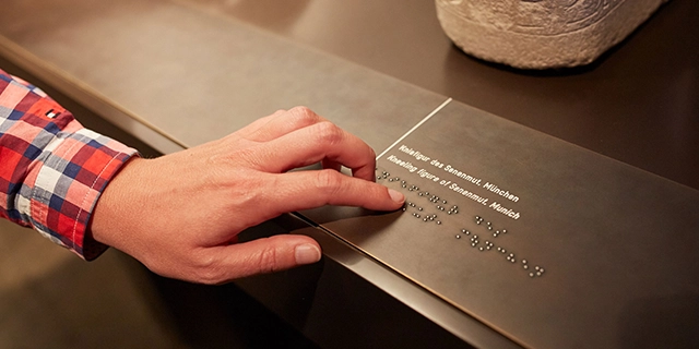 Museumsbesucherin vor einem Ausstellungsstück. Sie tastet die Erläuterung zum Austellungstück in Braille-Schrift ab.