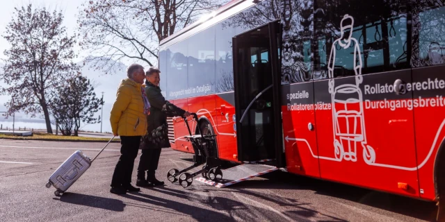 Einer Frau mit Rollator und einem Mann mit Koffer wird der Einstieg in den Bus mithilfe einer Rampe erleichtert. 