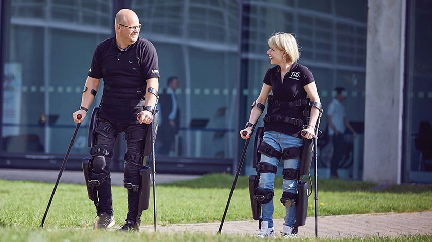 Ein Mann und eine Frau gehen mit ihren Exoskeletten und Gehhilfen spazieren.