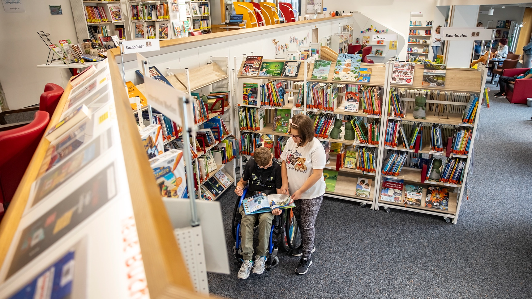 Ein Schüler im Rollstuhl blättert in der Schulbibliothek in einem Buch. Eine Schülerin steht neben ihm.