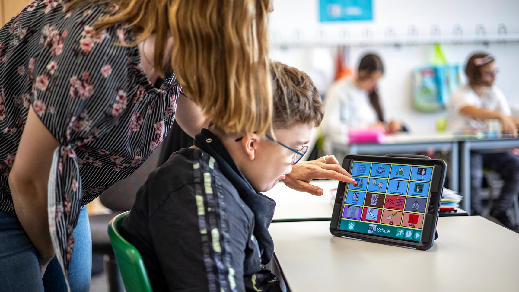 Ein Junge schaut auf ein Tablet in einem Klassenzimmer. Auf dem Bildschirm ist eine App als Kommunikationshilfe zu sehen. Eine Frau steht hinter dem Jungen und tippt auf den Bildschirm.