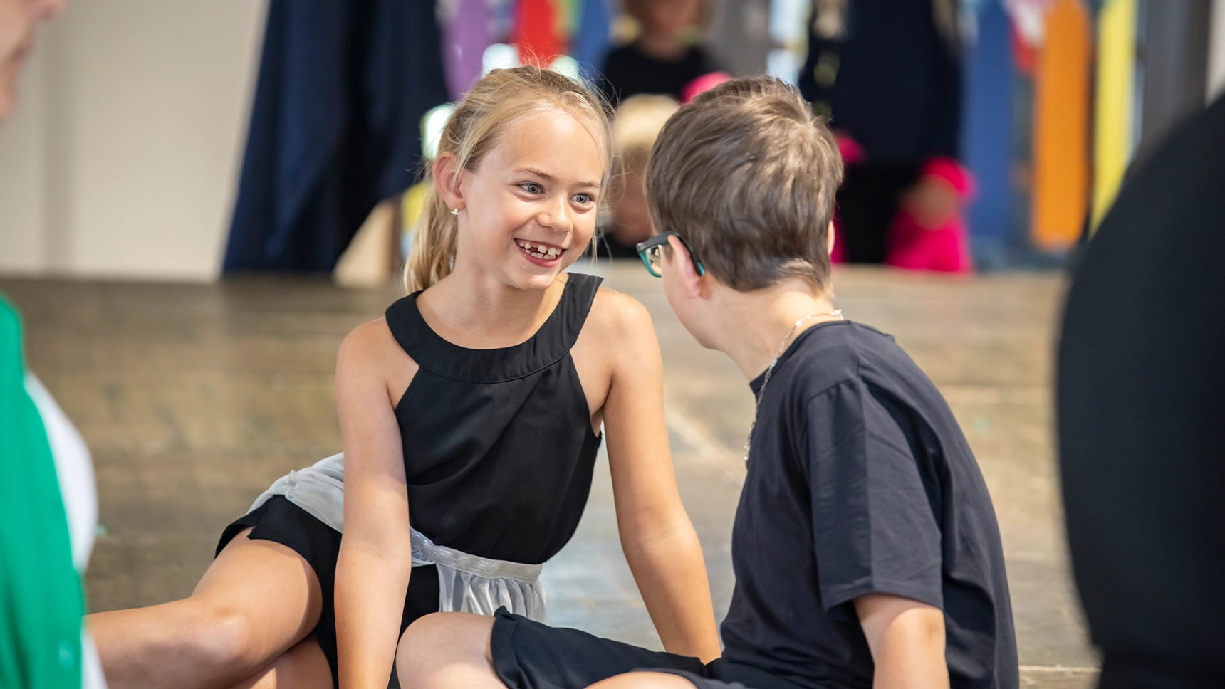 Ein Mädchen und ein Junge sitzen auf einer Theaterbühne. Das Mädchen lächelt den Jungen an.