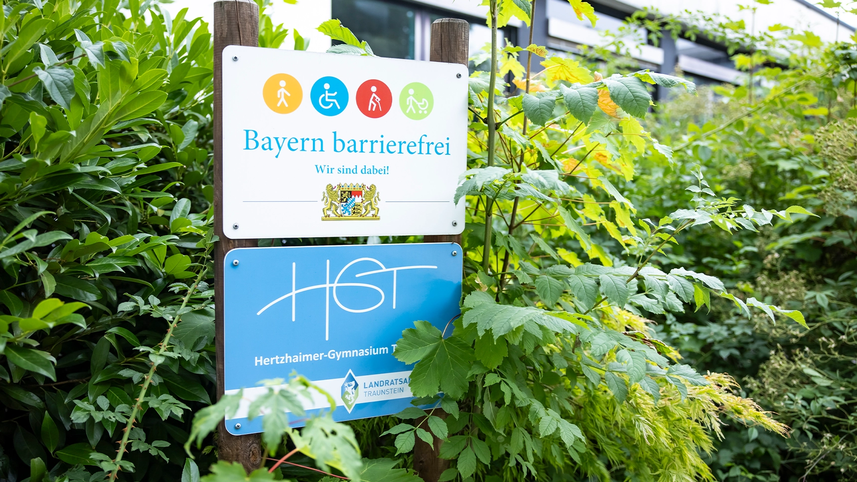 Schilder vor dem Schulgebäude zeigen das Signet „Bayern barrierefrei“ und das Logo des Hertzhaimer-Gymnasiums.