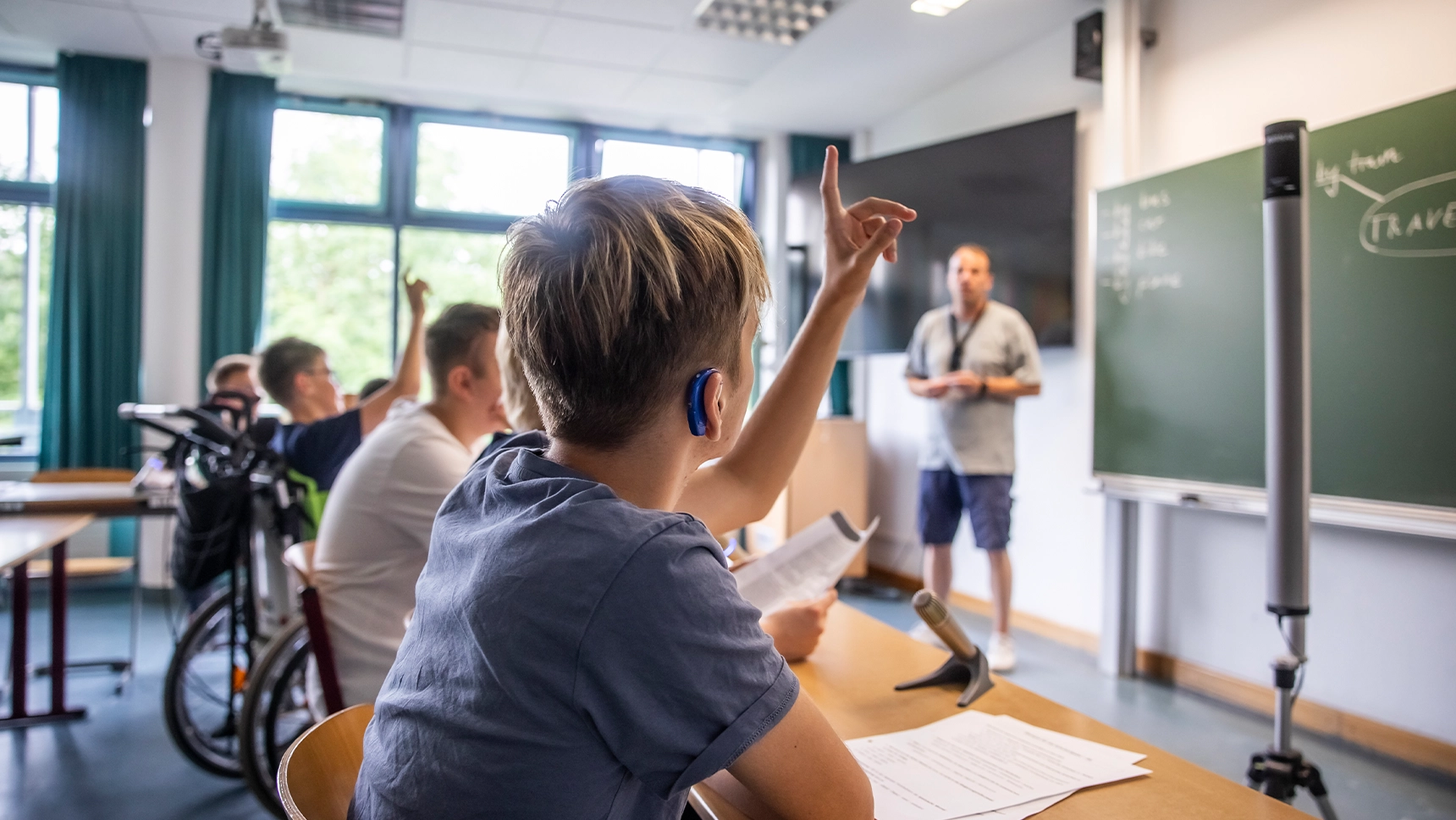 In einem Klassenraum mit Mikrofonen und Lautsprechern melden sich ein Schüler mit Hörgerät und ein Schüler mit Rollstuhl per Handzeichen zu Wort.