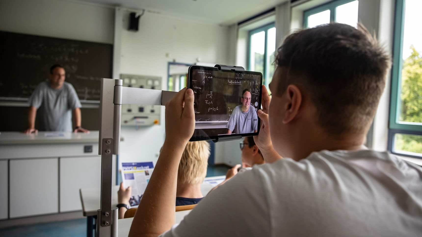 Junge im Schulunterricht mit einem Tablet in den Händen, auf dem der Lehrer am Pult nah zu sehen ist.