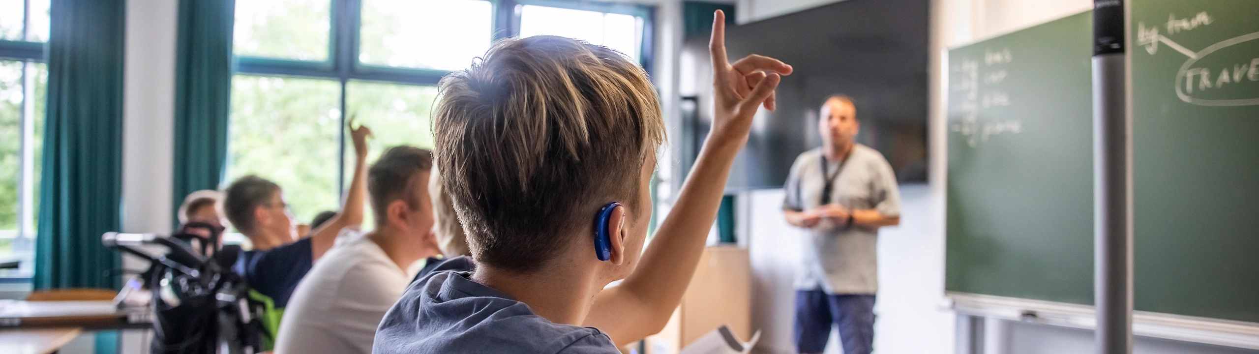 Ein Klassenzimmer: Ein Junge mit Hörgerät hebt die Hand.