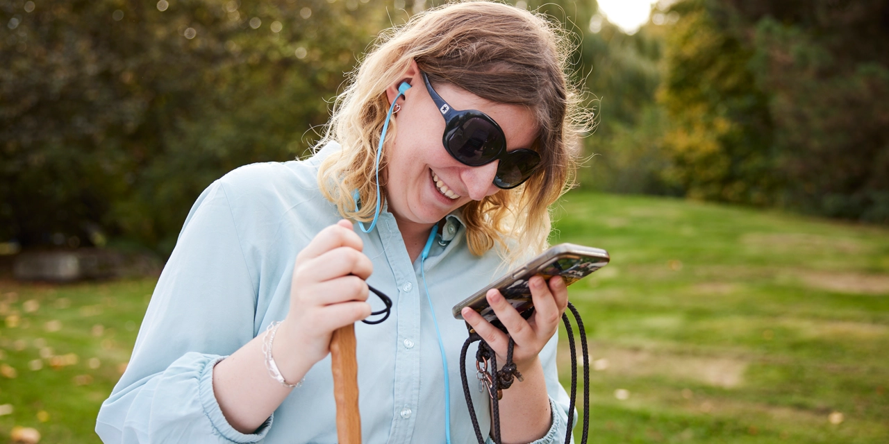 Eine Frau mit Sonnenbrille schaut lächelnd auf ihr Smartphone. 