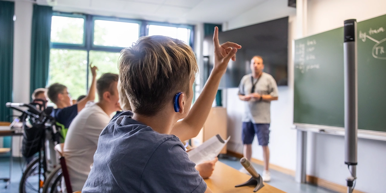 Ein Junge hebt im Schulunterricht die Hand. Er trägt ein Hörgerät. 