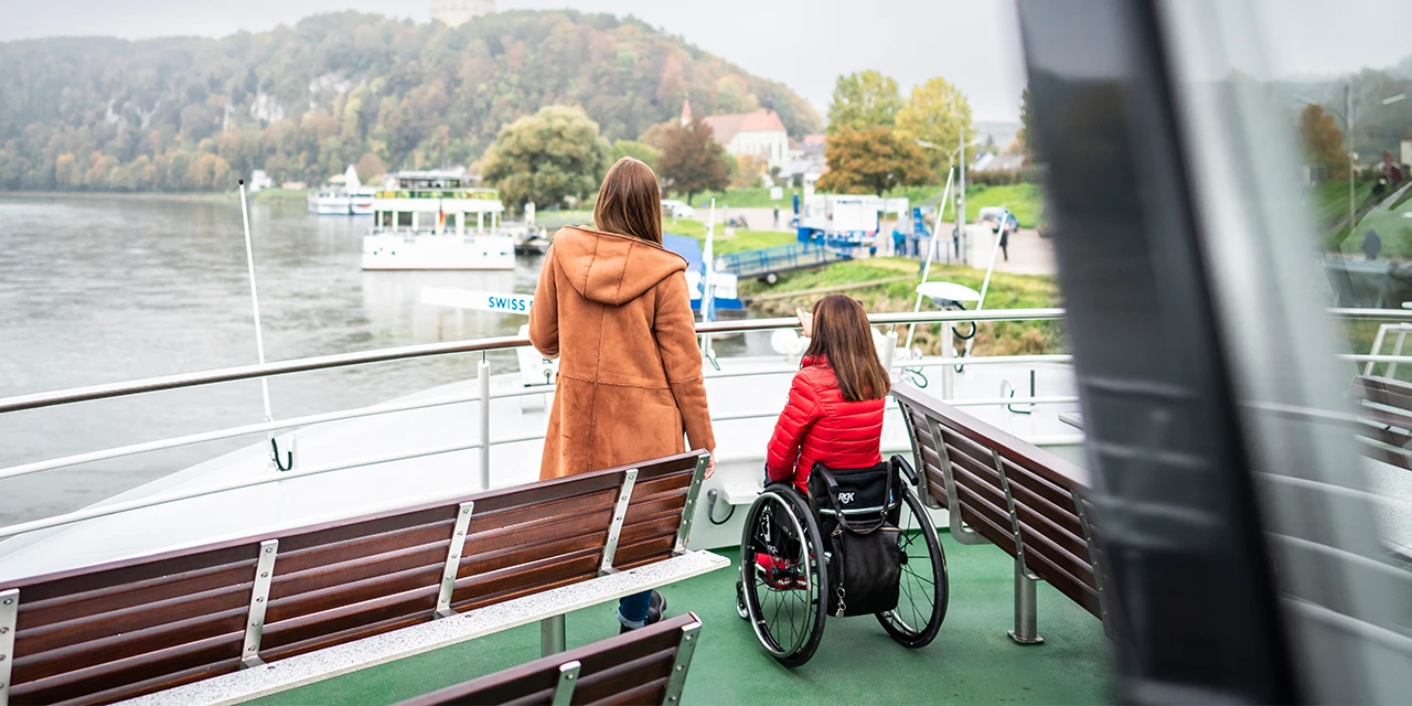 Eine Mutter steht mit ihrer Tochter im Rollstuhl auf einer Fähre.