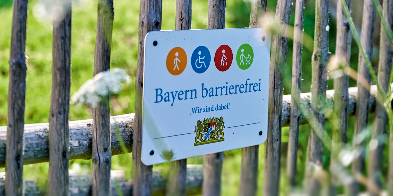 Das Signet „Bayern barrierefrei“ hängt an einem Holzzaun.