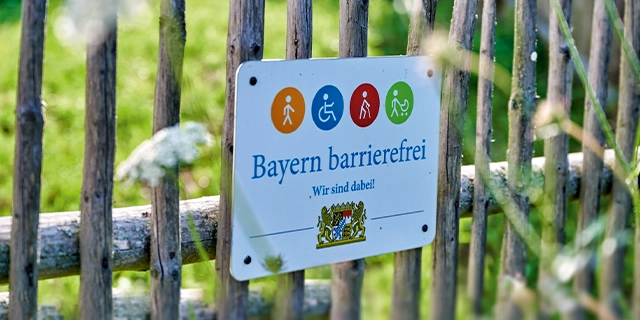 Das Signet „Bayern barrierefrei“ hängt an einem Holzzaun.